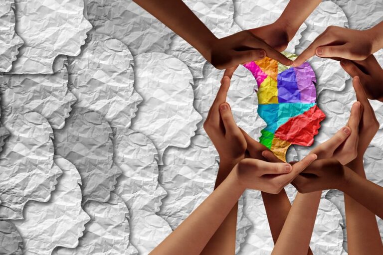 Hands of many different people reaching in to form a heart around a colorful paper silhouette of a head.